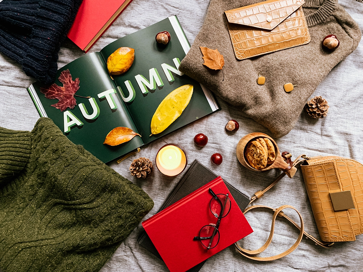 Flat lay with sweaters, accessories, books, and autumn leaves surrounding an open magazine, evoking cozy fall fashion inspiration.
