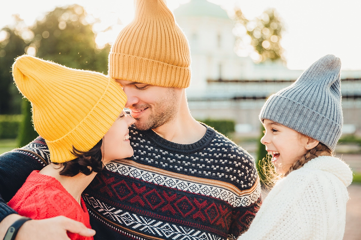 Family wearing colorful knit beanies and patterned sweaters, enjoying a sunny fall day together in warm seasonal clothing.