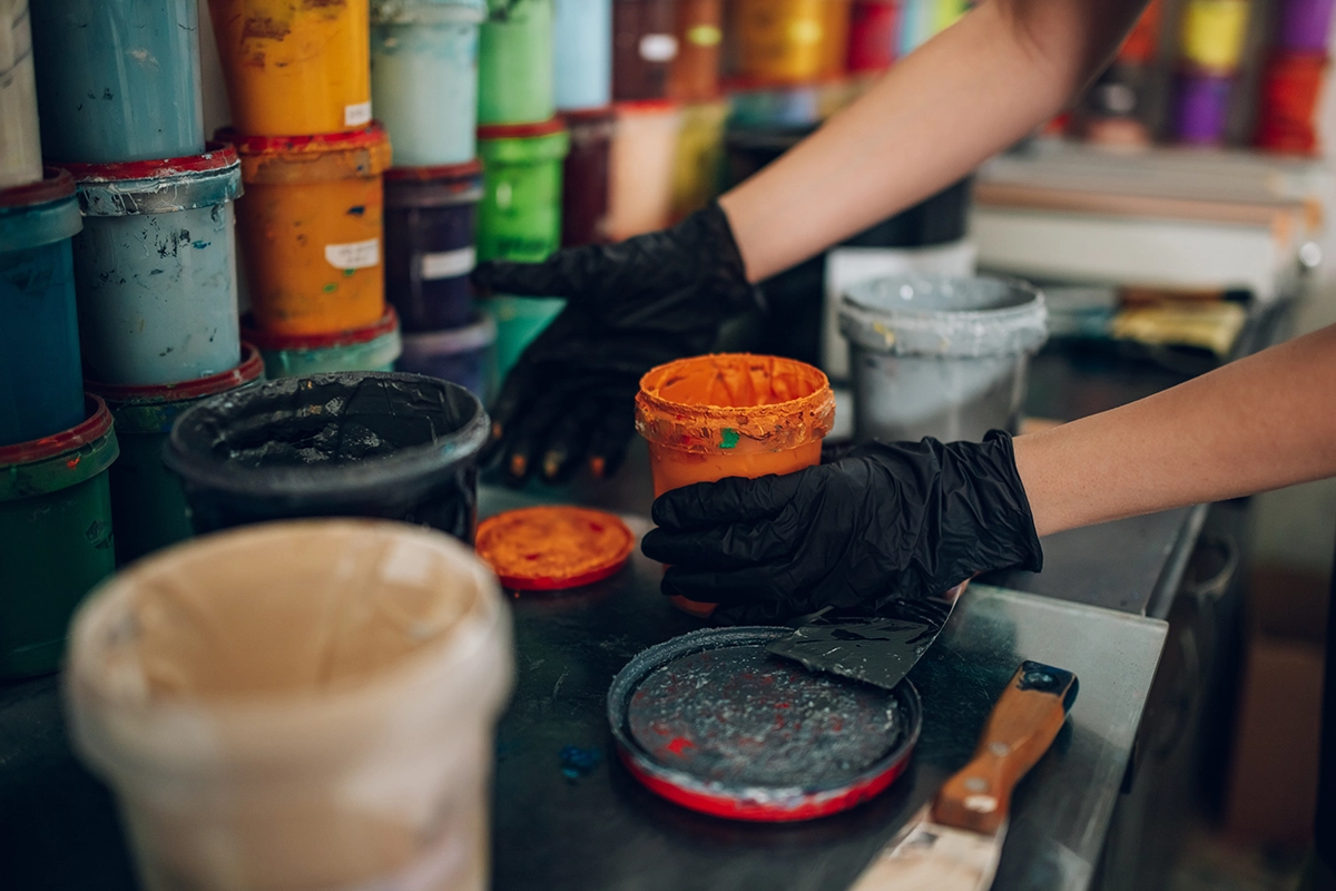 Hands in black gloves handling vibrant orange screen printing ink in a workshop surrounded by colorful paint containers.