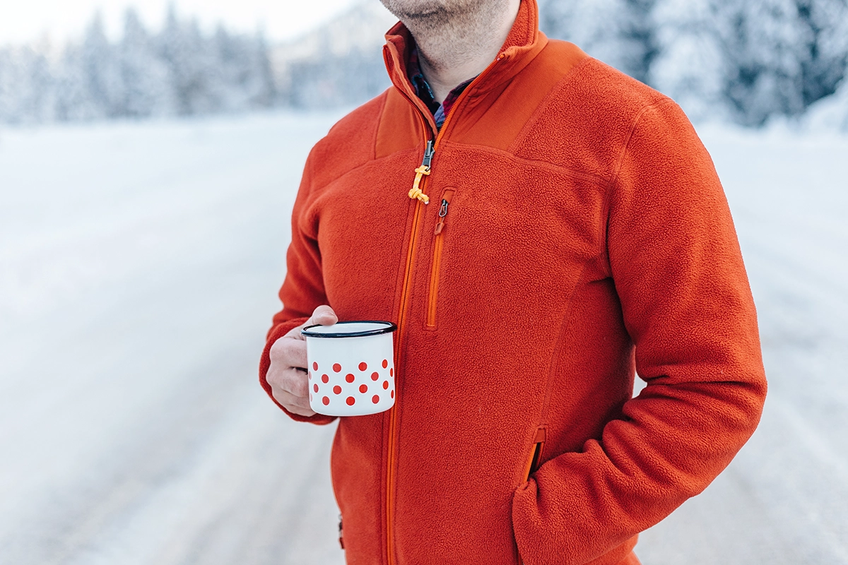 Person wearing a bright orange fleece jacket holding a coffee mug outdoors in a snowy Alaskan landscape, showcasing cozy fall apparel.