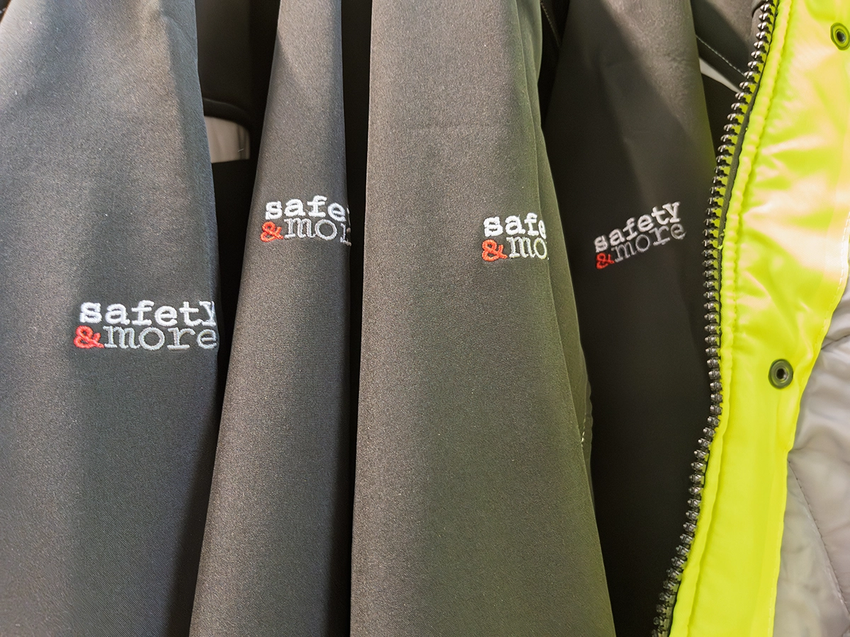 Black jackets embroidered with the logo “safety & more,” displayed on hangers beside a high-visibility yellow jacket.
