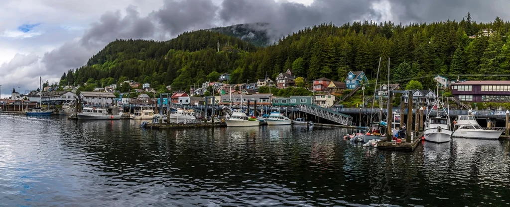 Panoramic view of Ketchikan, Alaska, showing colorful hillside homes, forested mountains, and fishing boats docked along the harbor under cloudy skies.
