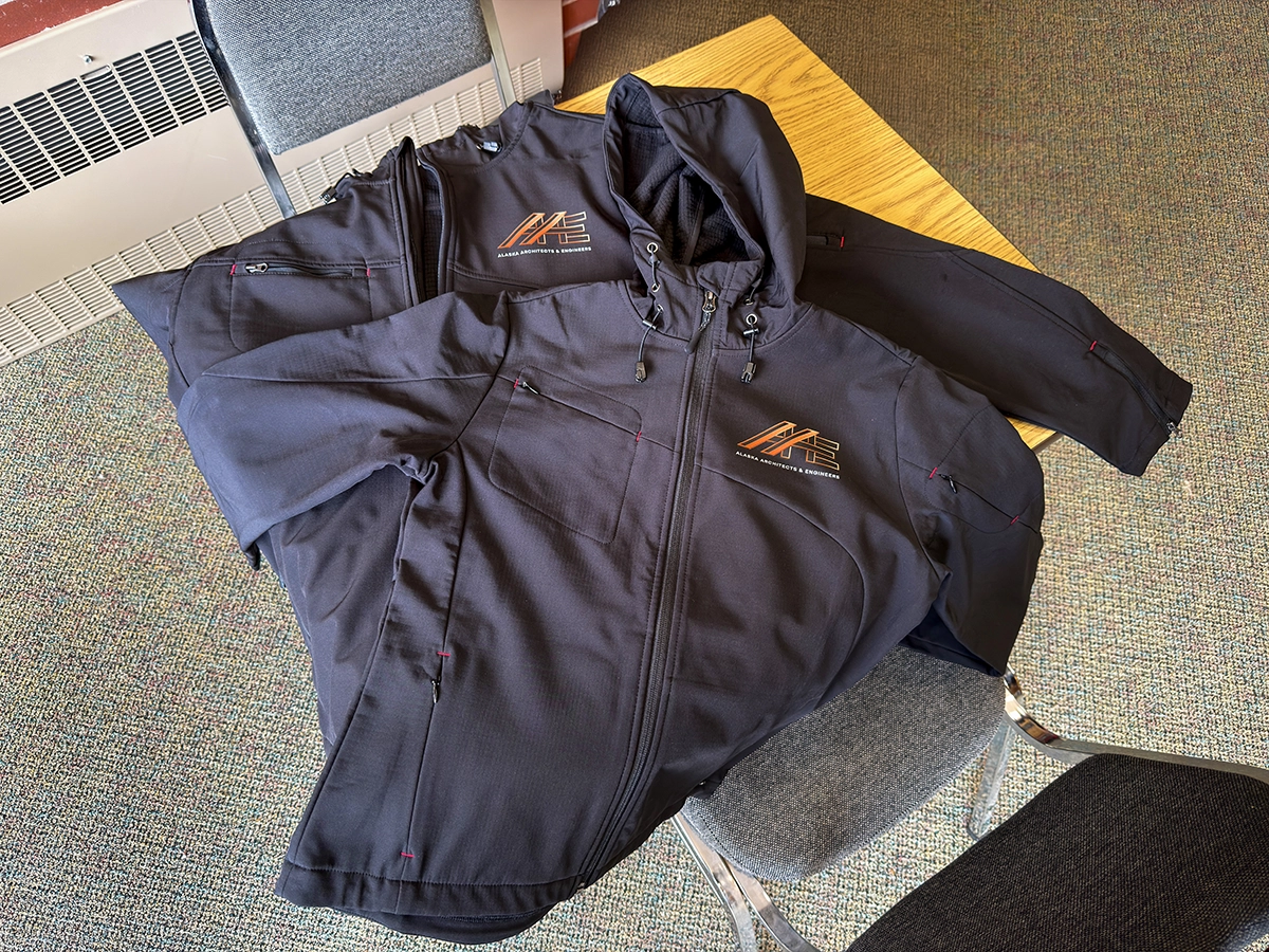 Black embroidered jackets with the Alaska Architects & Engineers logo folded neatly on a chair inside an office space.