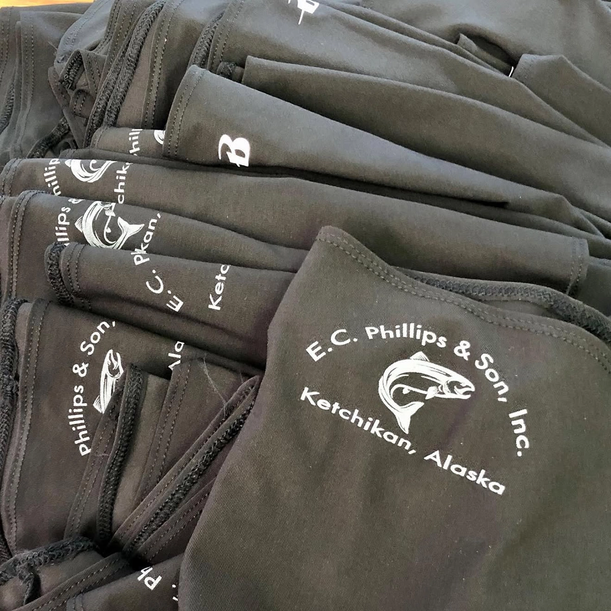 Stack of black fabric masks printed with the logo “E.C. Phillips & Son, Inc. Ketchikan, Alaska” and a fish graphic.