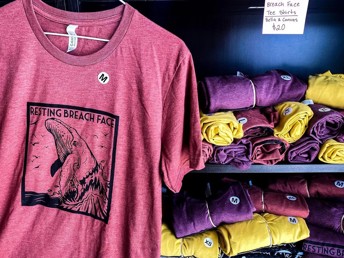 Display of folded maroon, yellow, and purple T-shirts with one shirt hanging in front reading “Resting Breach Face” with a whale graphic design.