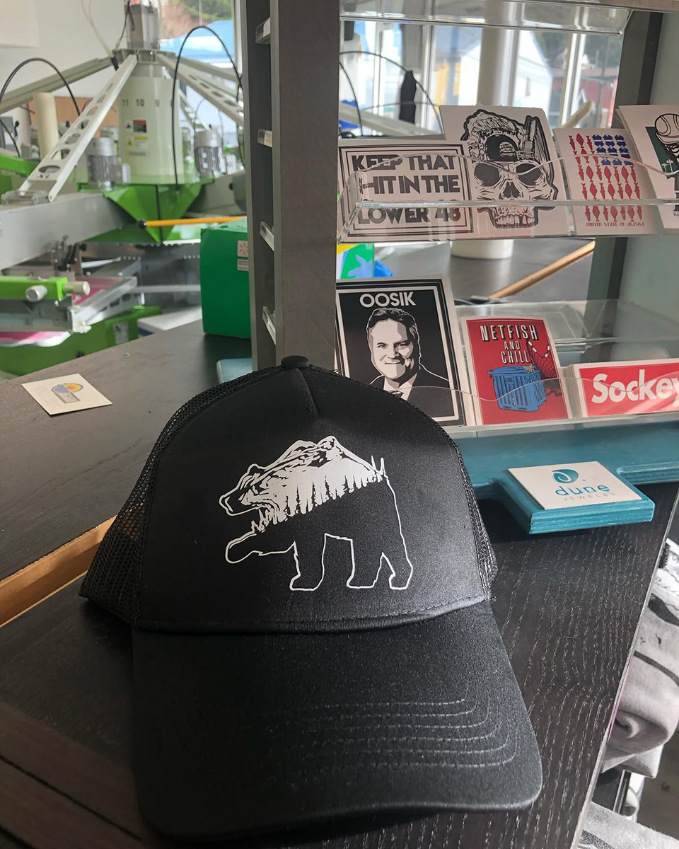 Black trucker hat with a white bear and tree silhouette design displayed on a counter in front of a sticker rack and screen-printing equipment.