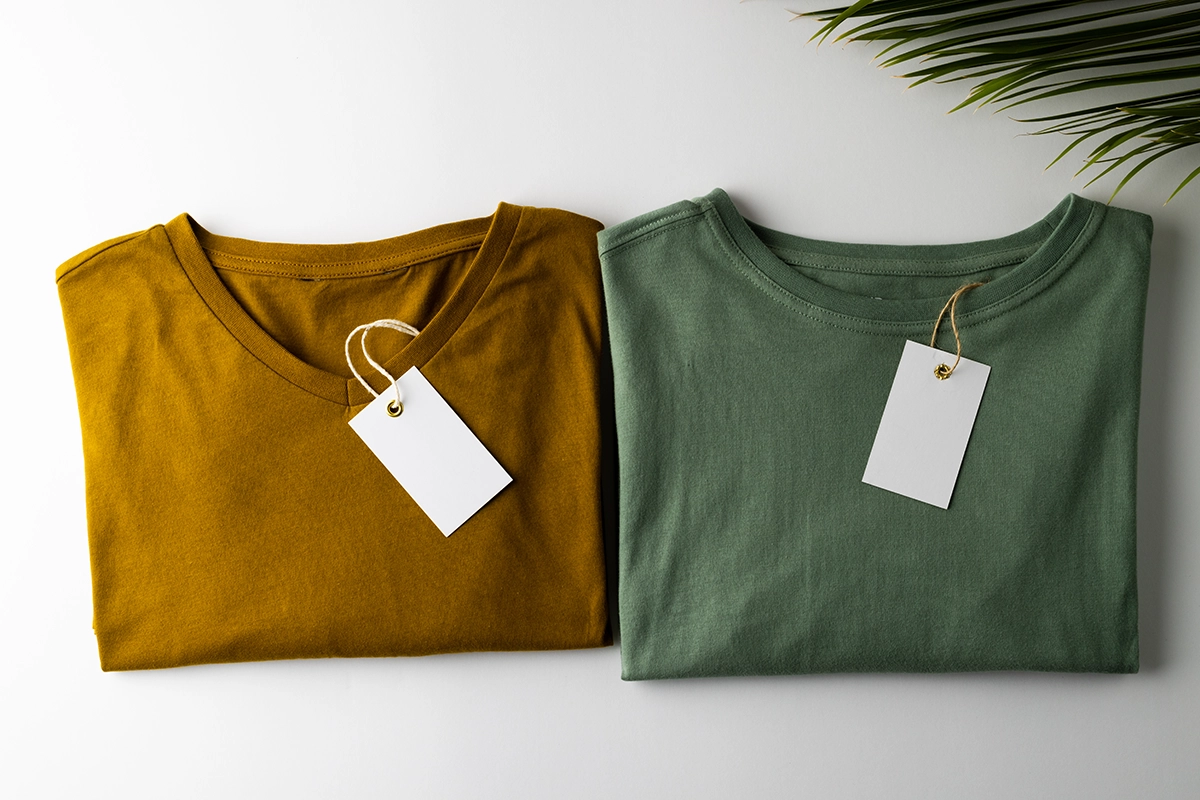 wo folded t-shirts in mustard yellow and sage green with blank white swing tags, styled on a white surface with palm leaves for a modern look.