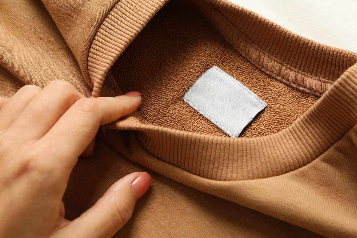 Close-up of a hand lifting the collar of a tan sweatshirt to reveal a blank label inside the soft terry fabric
