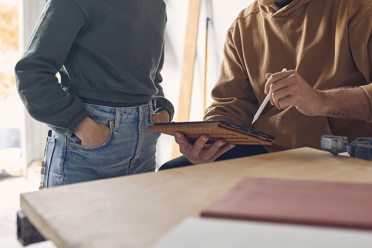Two people wearing casual sweatshirts in a workshop setting reviewing designs on a tablet together at a wooden table.