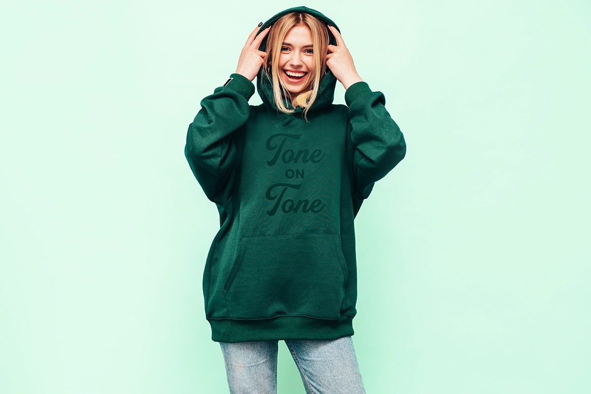 Smiling woman wearing a dark green hoodie with “Tone on Tone” printed on the front, posing confidently against a mint background.