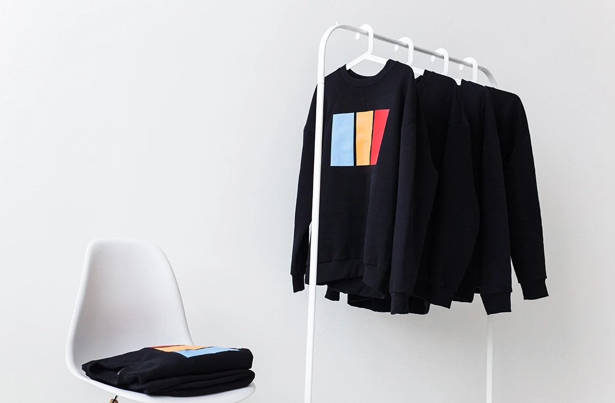 Black sweatshirts with colorful geometric prints hanging on a white rack beside a white chair, arranged for a clean product display.