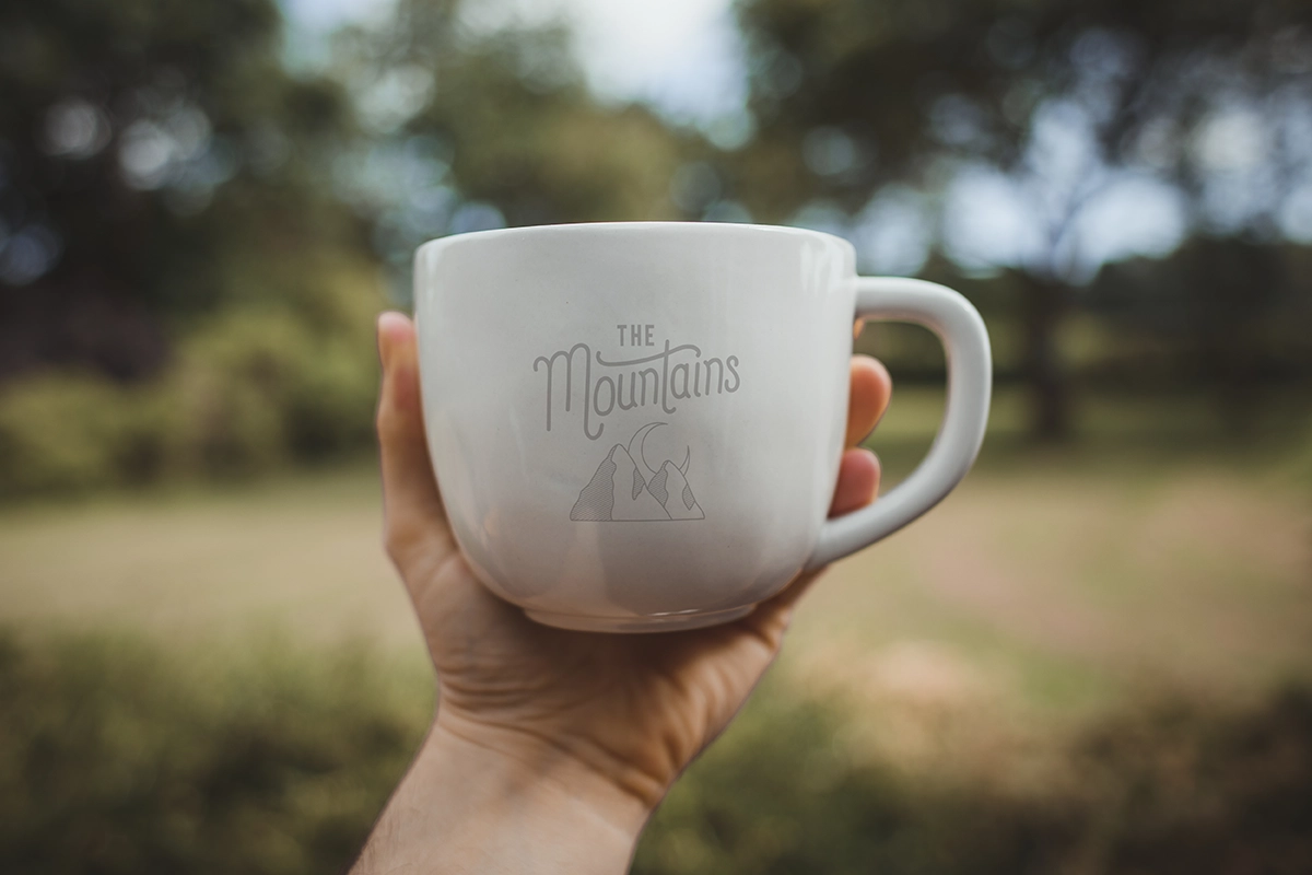 Hand holding a white ceramic mug with “The Mountains” logo engraved on the side, set against an outdoor blurred background.
