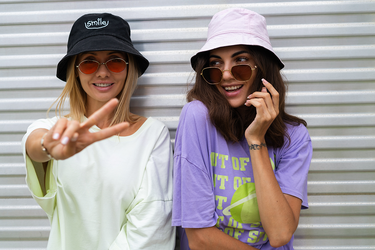 Two women posing playfully in casual streetwear; one wearing a black bucket hat with “smile” embroidery and the other in a lilac T-shirt and pink bucket hat, both in sunglasses.