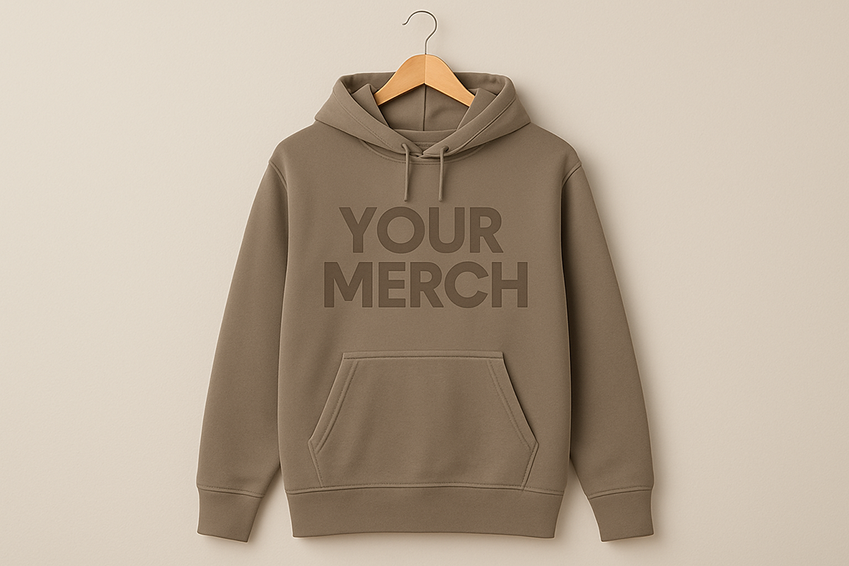 Tan hoodie hanging on a wooden hanger against a neutral wall with the text “Your Merch” printed on the front in matching tone.
