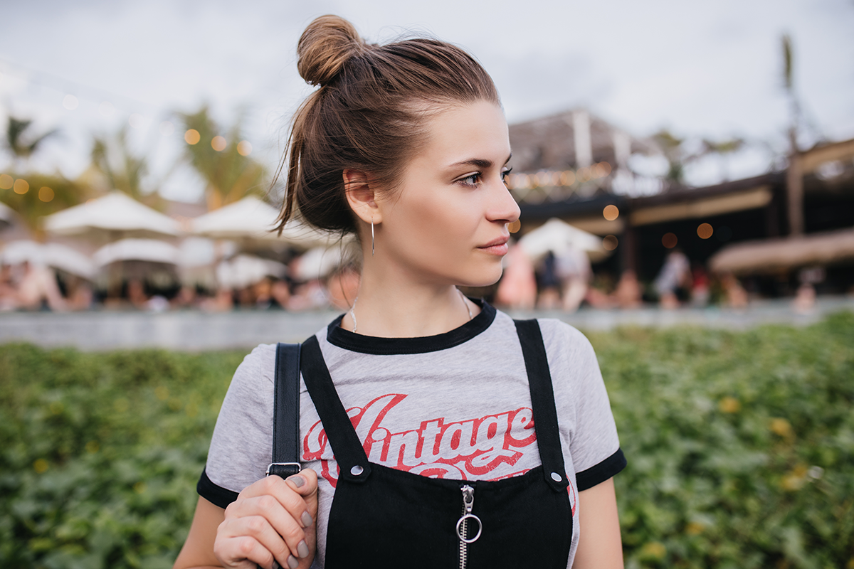 Woman outdoors wearing a gray ringer T-shirt with red vintage lettering and black overalls, looking to the side with a casual expression.