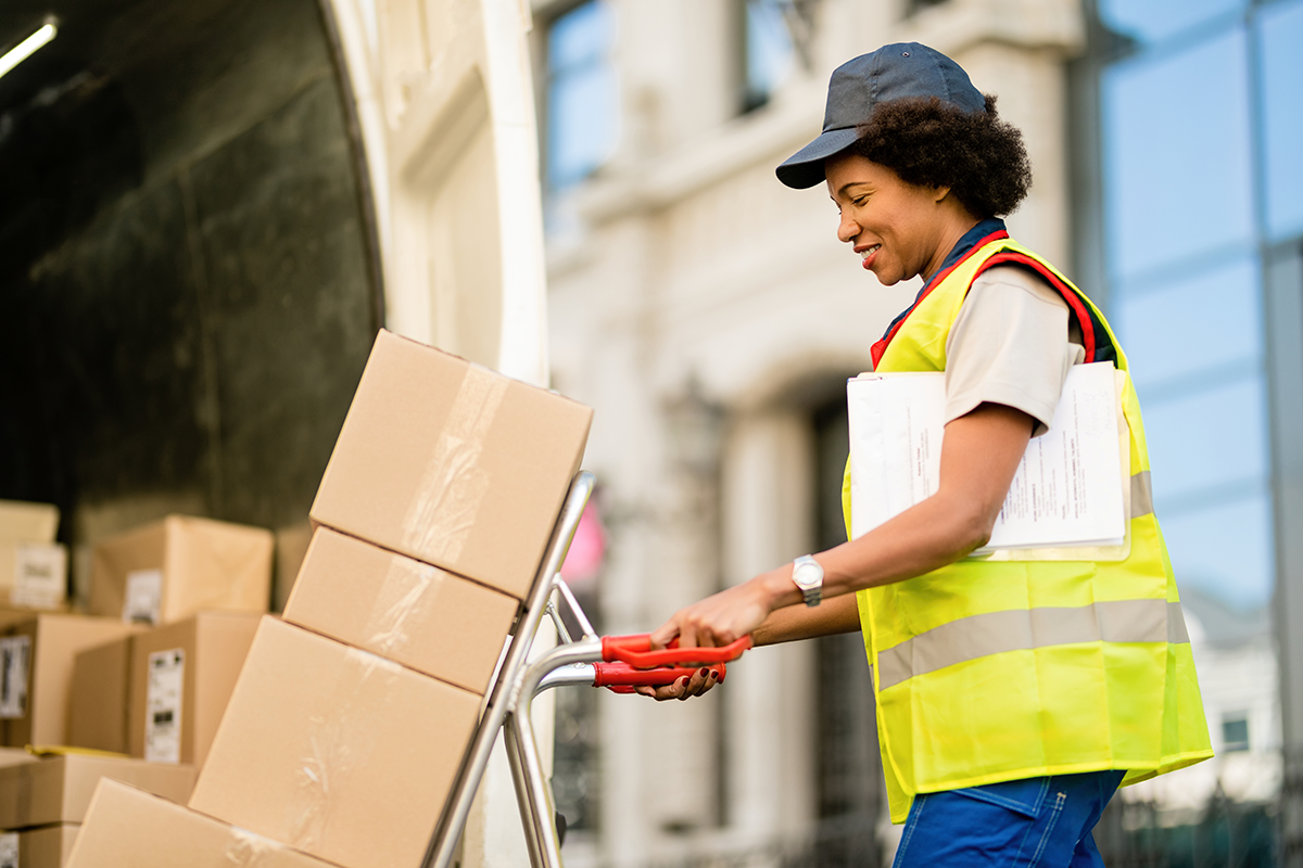Delivery worker pushing hand truck with stacked cardboard boxes outside commercial building