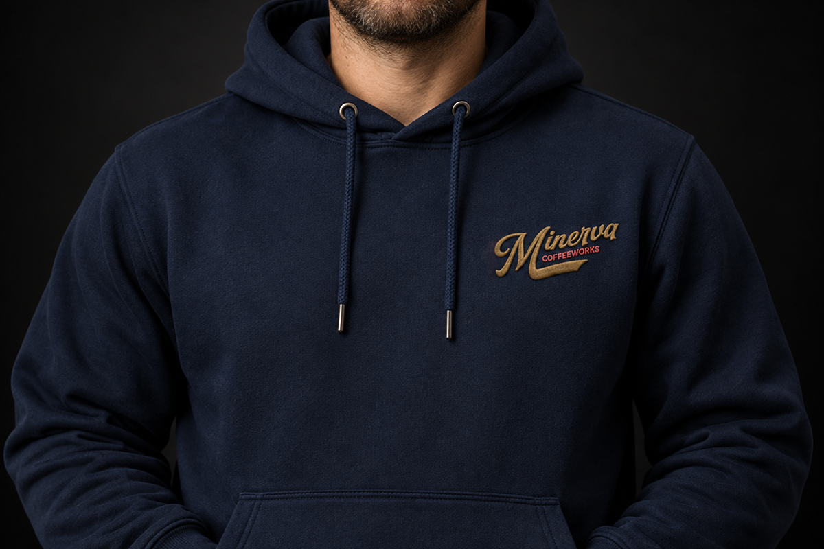 Close-up of a person wearing a navy hoodie with a gold and red embroidered “Minerva Coffeeworks” logo on the chest.