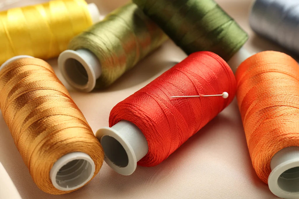 Close-up of embroidery thread spools in warm fall colors, including gold, green, red, and orange, with a sewing needle resting across the red spool.