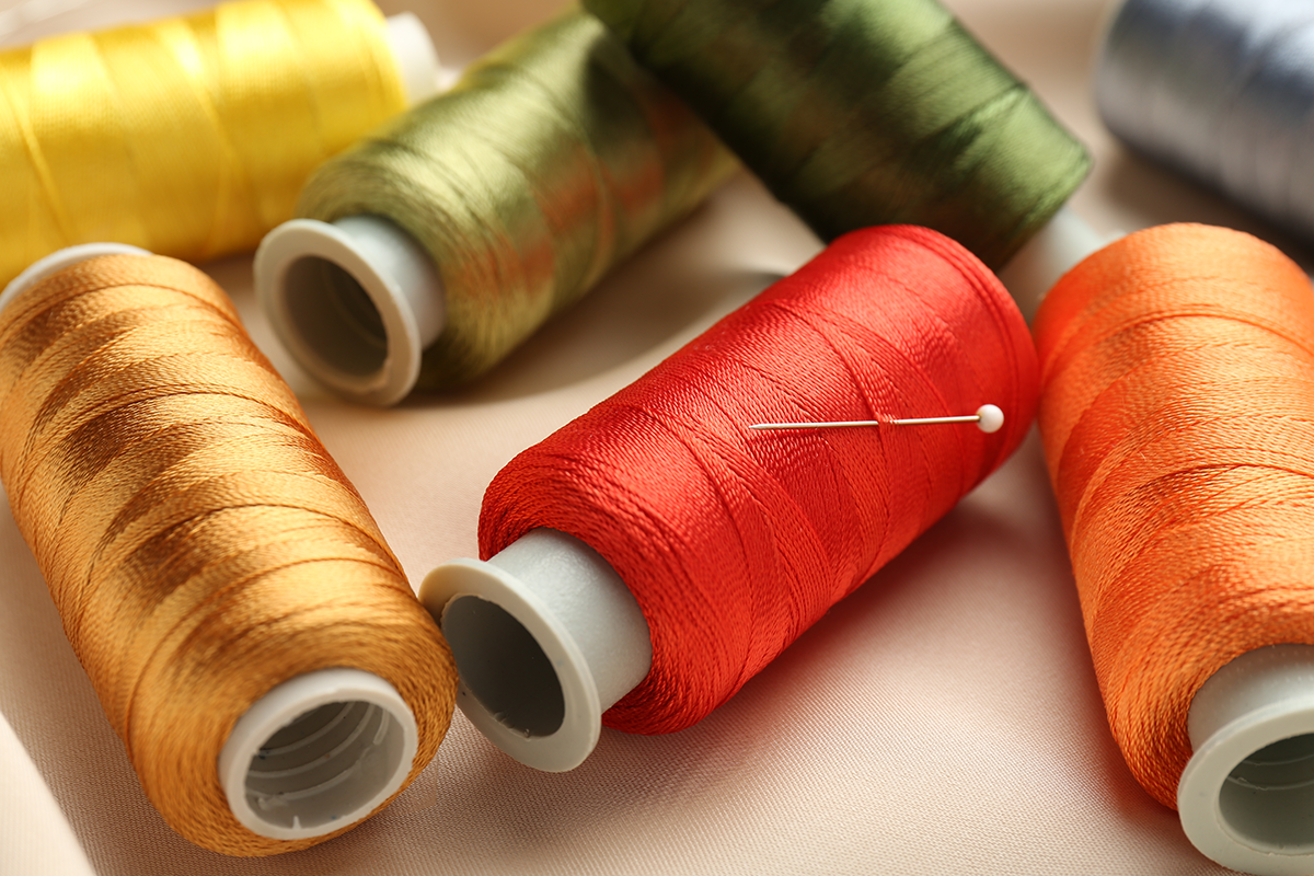 Close-up of embroidery thread spools in warm fall colors, including gold, green, red, and orange, with a sewing needle resting across the red spool.
